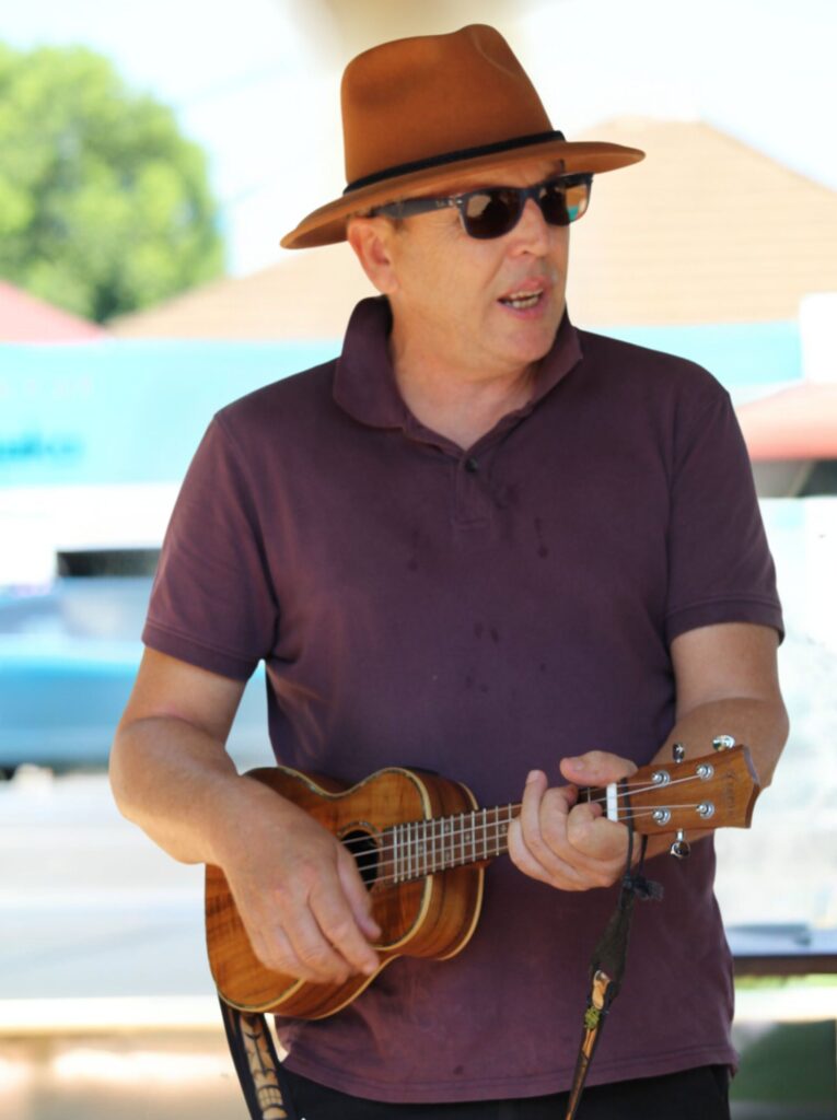 Stephen Ewart playing ukulele with a calm, serious expression. His posture and focus reflect his decades of musical experience and dedication.